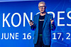 Man in a blazer speaking on stage against a blue conference backdrop.