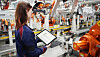 Worker using a tablet in a factory with robotic machinery and metal assembly equipment.