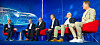 Five men seated on red chairs in front of a blue digital backdrop at a conference panel.