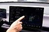 Hand pointing at a car infotainment touchscreen showing vehicle settings and a car image.