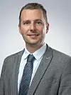 Man in a grey suit and blue tie posed against a plain grey studio backdrop.