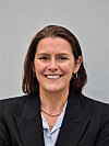Smiling woman in a black blazer and striped shirt against a grey background.