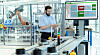 Man using a tablet next to a factory conveyor line and a control screen.