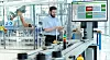 Man using a tablet next to a factory conveyor line and a control screen.