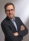 Portrait of a man in glasses wearing a dark blazer with his arms folded.