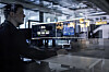 Man seated at a desk in a glass-walled office looking at several computer monitors.