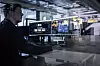 Man seated at a desk in a glass-walled office looking at several computer monitors.