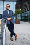 Man in a blue blazer standing beside a metal railing outside a modern office building.