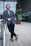 Man in a blue blazer standing beside a metal railing outside a modern office building.