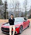Man standing beside a red Mercedes-Benz sports car on a roadside with trees and mountains behind.